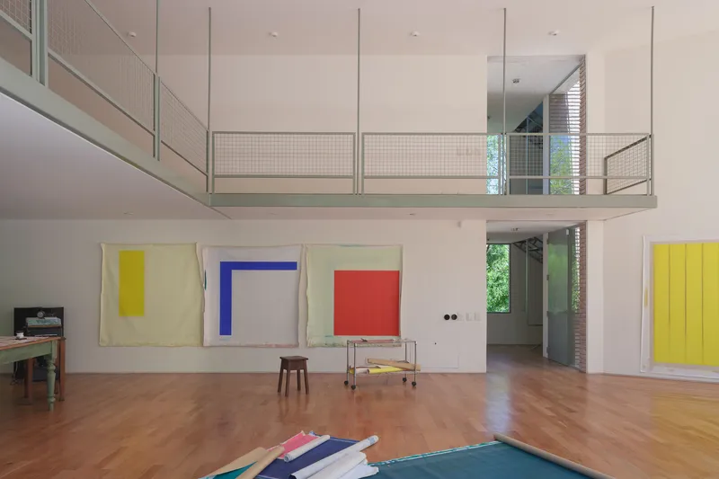 View across parquet floor toward mezzanine with wire mesh balustrade and doorway framing green foliage beyond