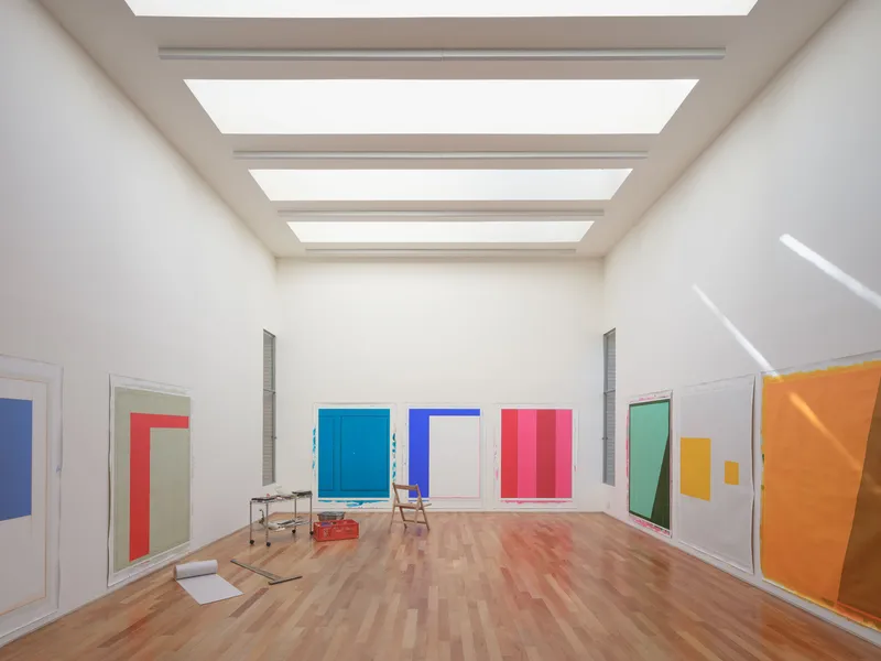 Gallery room with four parallel skylights casting even light across parquet floor and canvases displaying colored rectangles