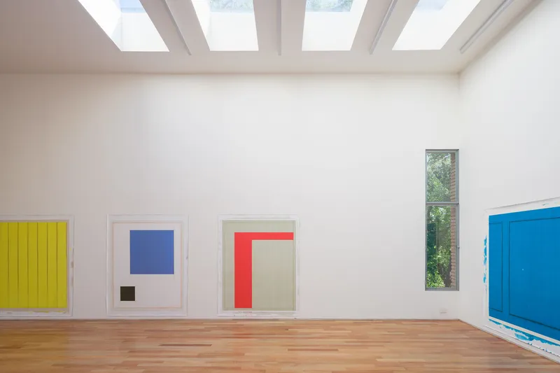 White gallery room with timber floor, geometric skylight beams, and tall vertical window overlooking trees