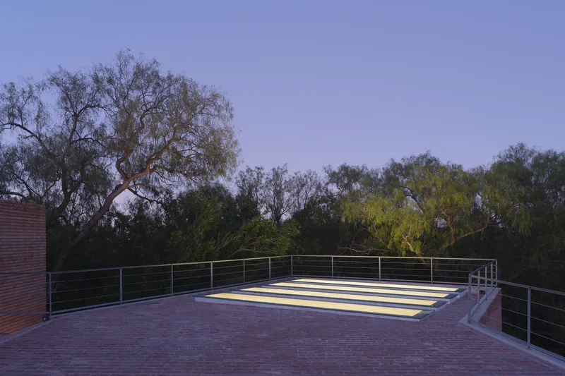 Rooftop terrace with horizontal brick paving, cable railing, and illuminated skylight panel at dusk
