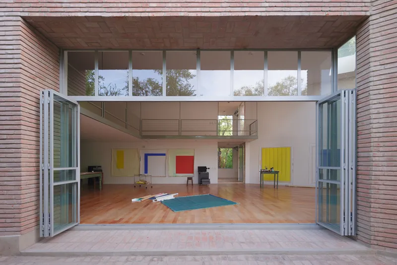 Open steel-framed glass doors revealing a double-height studio space with colorful canvases and wood flooring