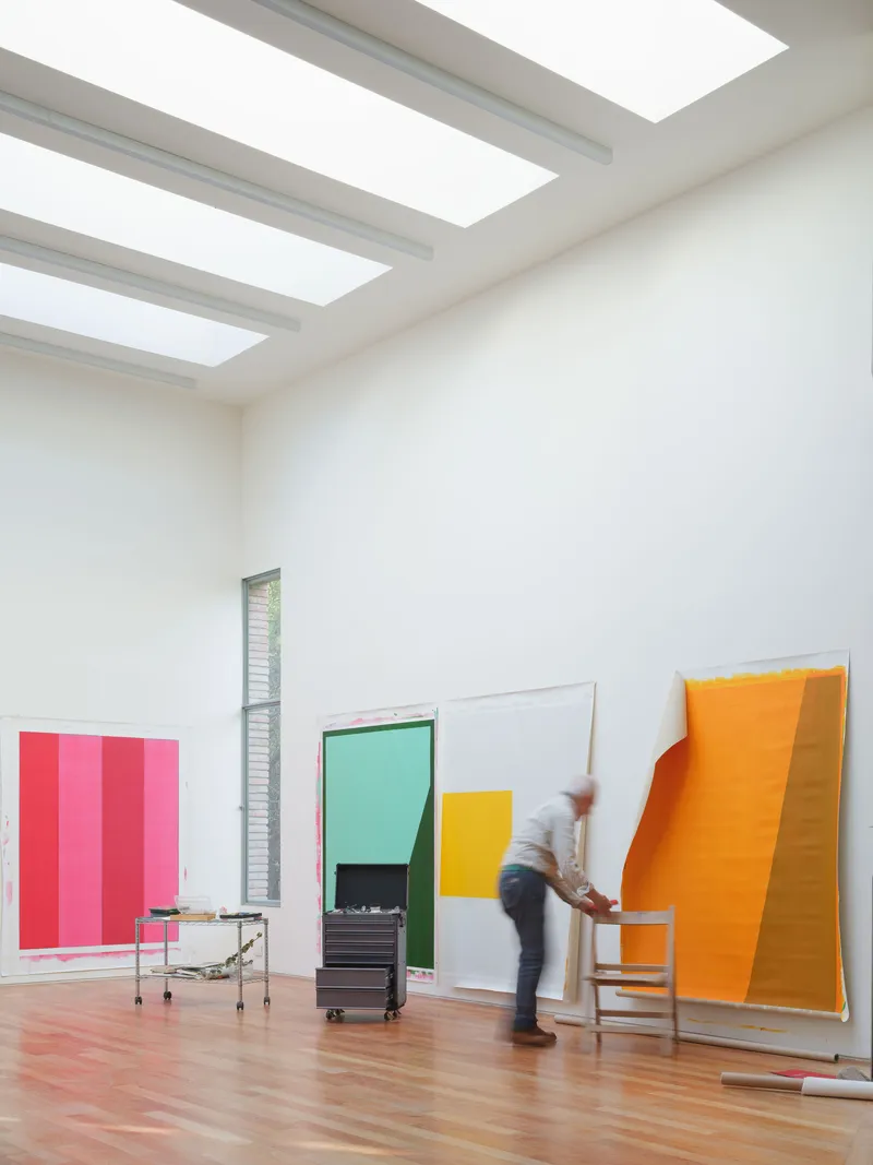 Gallery space with skylights illuminating a person moving past leaning canvases on polished wood floors