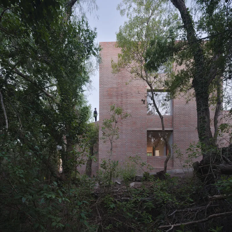 Three-story brick volume with recessed entry and figure on rooftop terrace framed by mature trees
