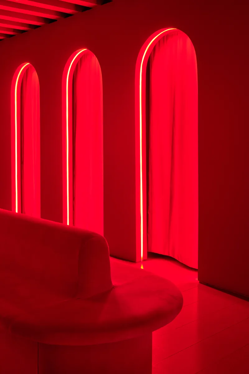 Three red neon-framed arched openings with draped fabric and a circular upholstered seat
