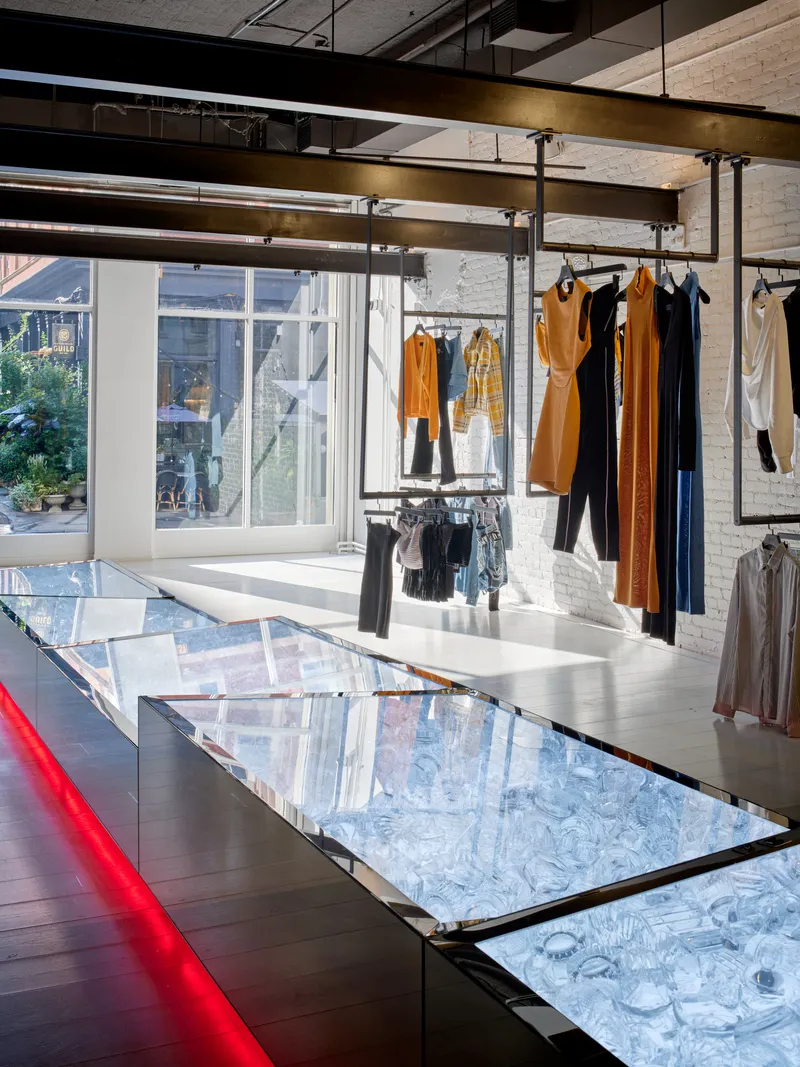 Glass display table with blue illuminated base beneath suspended clothing racks and brass ceiling beams