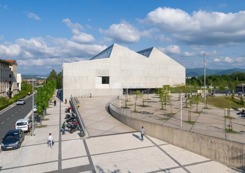 Pale timber-clad facade with peaked roofline rising above a public plaza with young trees and pedestrians