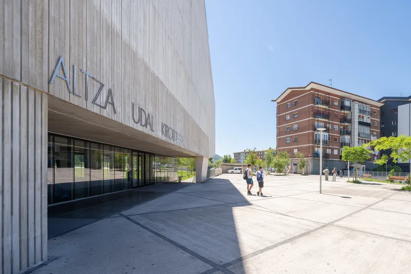 Vertically ribbed concrete facade with recessed entrance and pedestrians crossing the plaza under blue sky