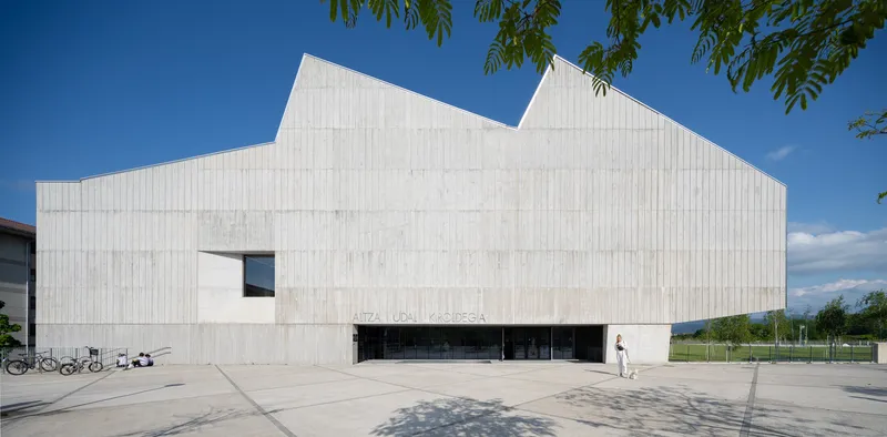 Ribbed concrete facade with angular roofline and horizontal entrance slot beneath tree branches