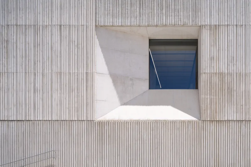 Vertical ribbed concrete facade with a recessed window opening revealing angled blue panels in bright sunlight