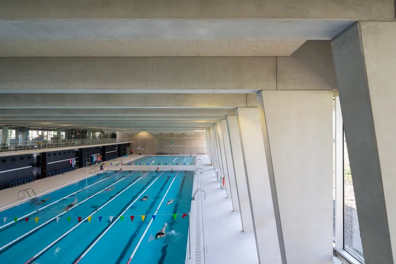 Interior view of the lap pool with angled concrete ceiling beams and swimmers in lanes