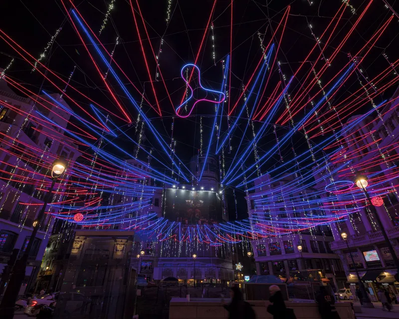Wide view of LED light installation radiating from central pillar over pedestrians in an urban plaza