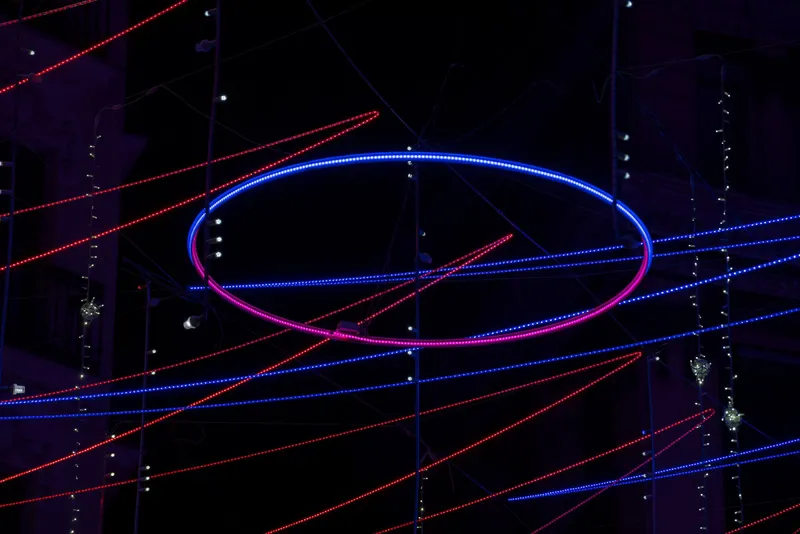 Illuminated elliptical ring with blue and pink LED lights suspended among horizontal light strands at night