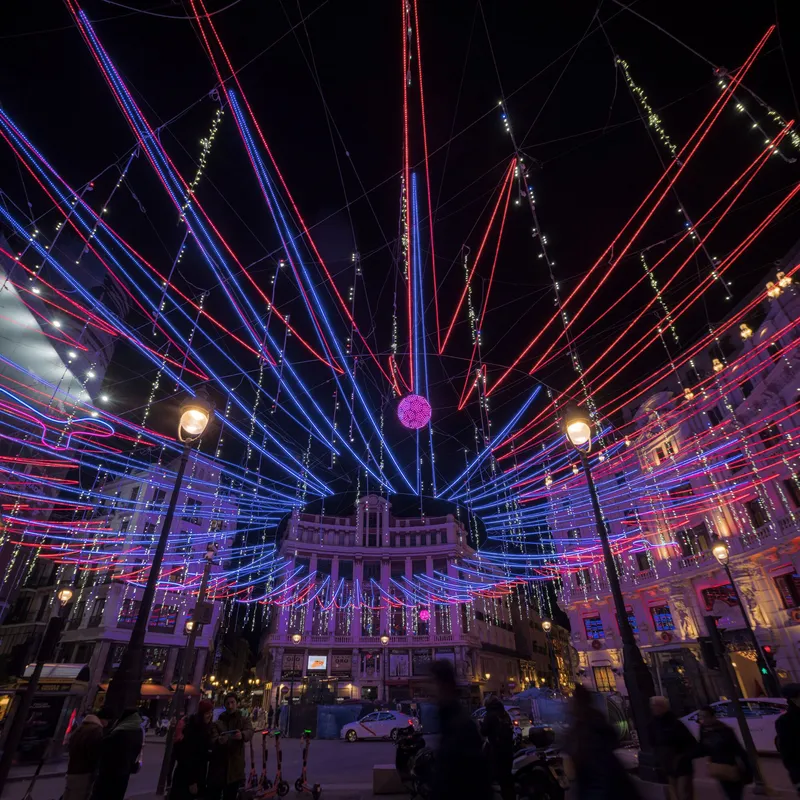 Radial LED installation converging above plaza with pedestrians and street lamps visible at dusk