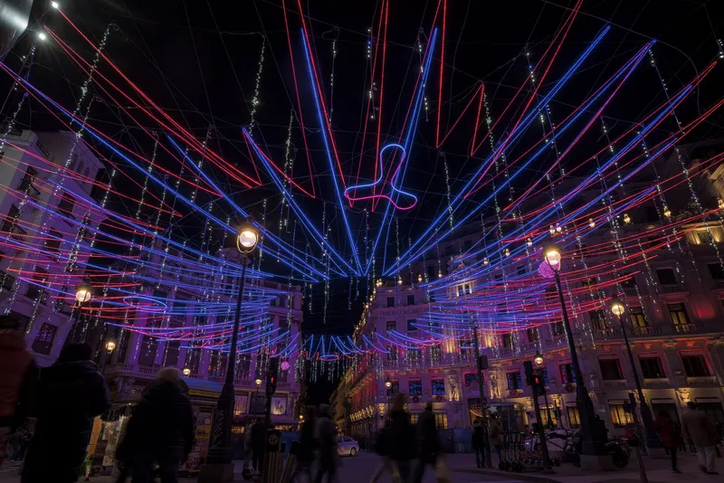 Overhead canopy of illuminated red and blue LED tubes radiating from a central point above a public square at night