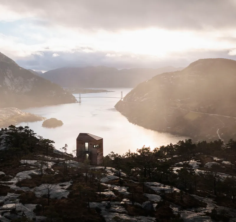 Timber cabin on a hillside overlooking a misty fjord with a suspension bridge at sunrise