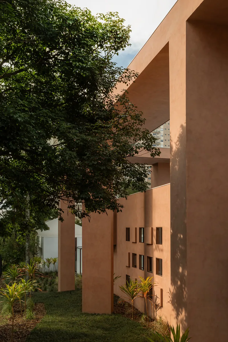 Terracotta stucco facade with scattered square windows beside mature trees casting dappled shadows