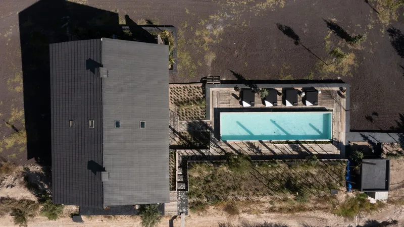 Aerial view of the rectangular volume with corrugated metal roof and adjacent pool deck surrounded by sandy vegetation