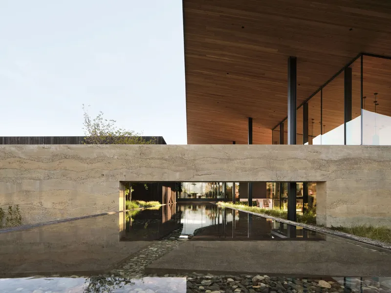 Rammed earth wall bridging a reflecting pool beneath timber soffit with steel columns at twilight