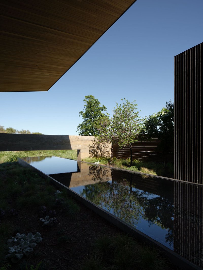 Reflecting pool edged with vertical black slats beneath a timber soffit under clear blue sky