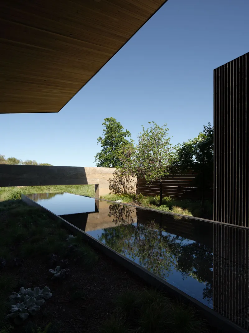 Reflecting pool edged with vertical black slats beneath a timber soffit under clear blue sky