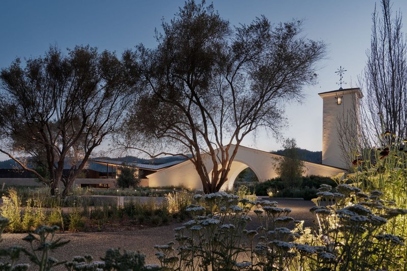 Arched facade and bell tower rising above a gravel landscape with mature trees at twilight