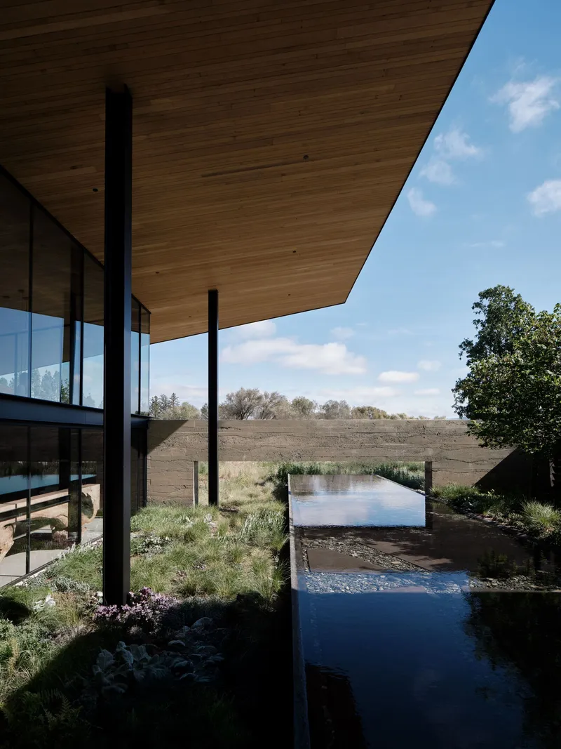 Overhanging timber soffit above a narrow water channel bordered by grasses and native plantings