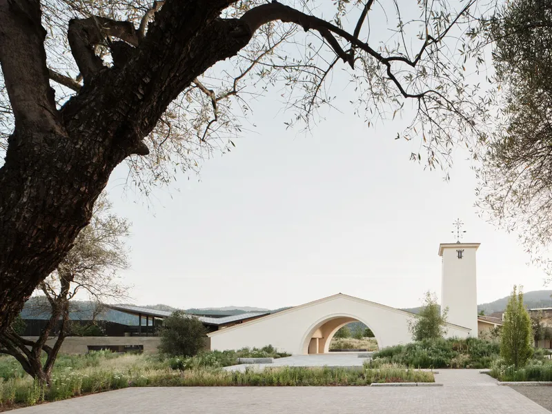 White stucco building with arched opening and bell tower framed by overhanging olive tree branches