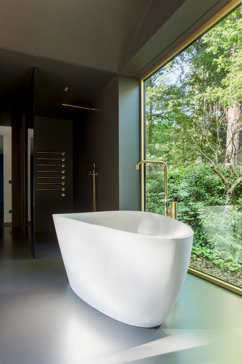 White sculptural bathtub with brass fittings on grey floor beside full-height glass overlooking dense foliage