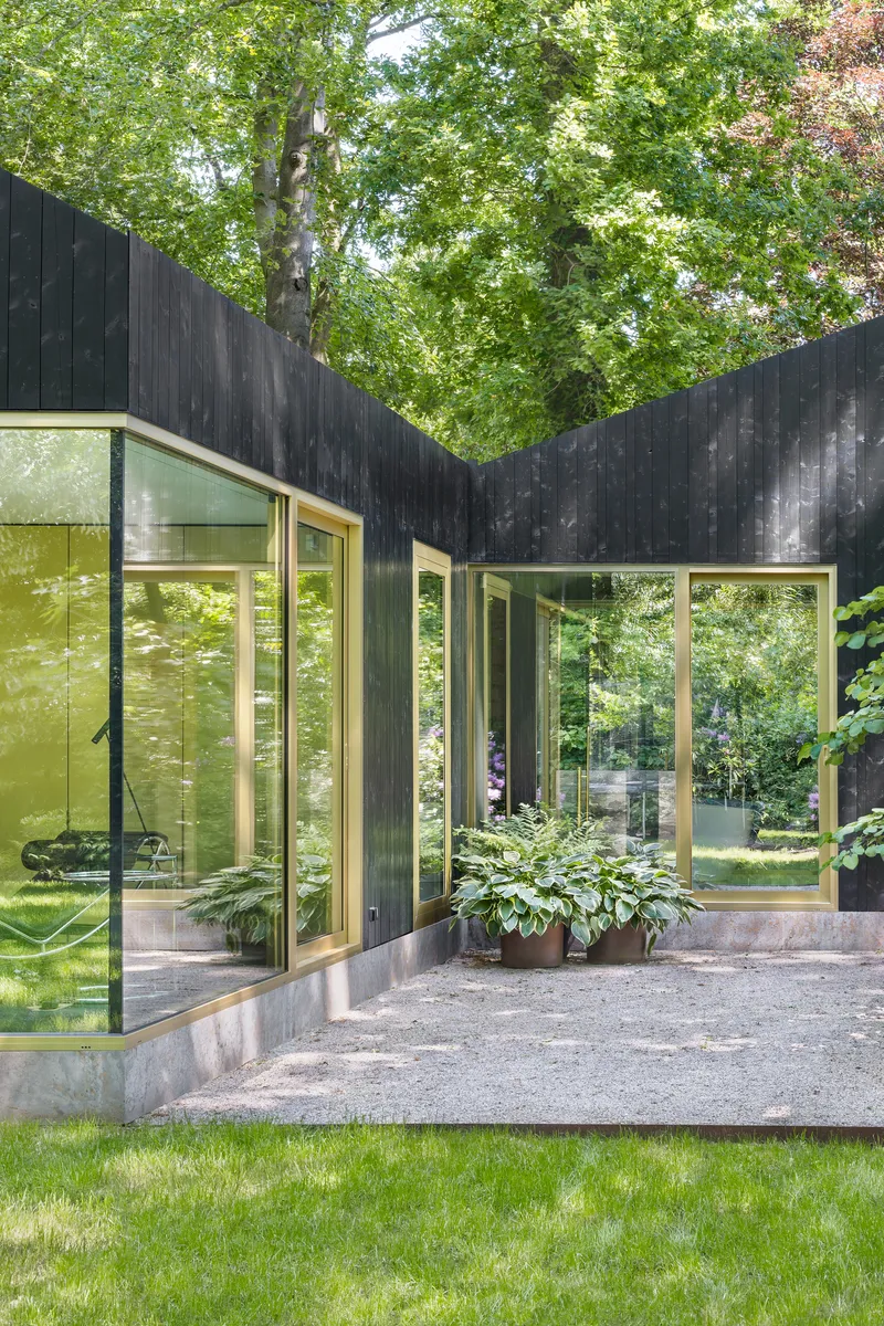 Corner junction of glass-walled rooms with concrete plinth and planted bed beneath dappled forest canopy