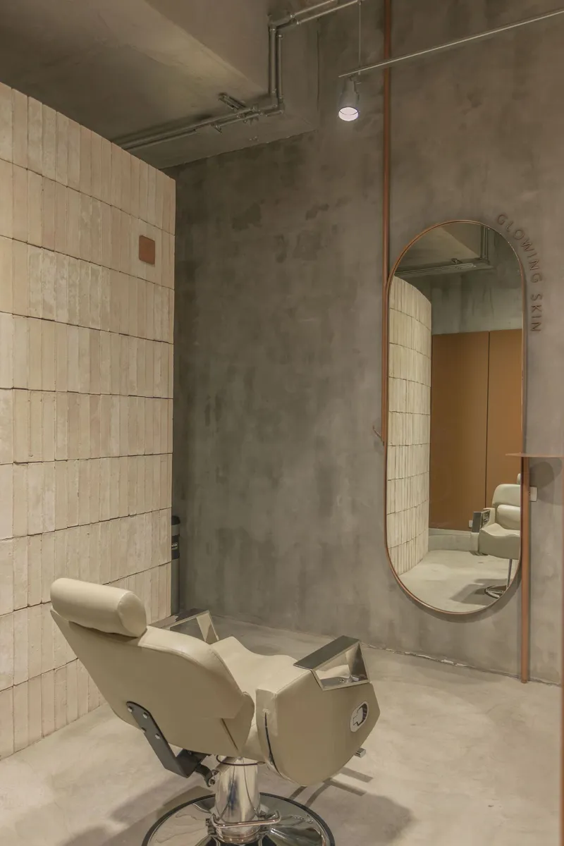 Salon chair beneath oval mirror framed in copper against polished concrete wall