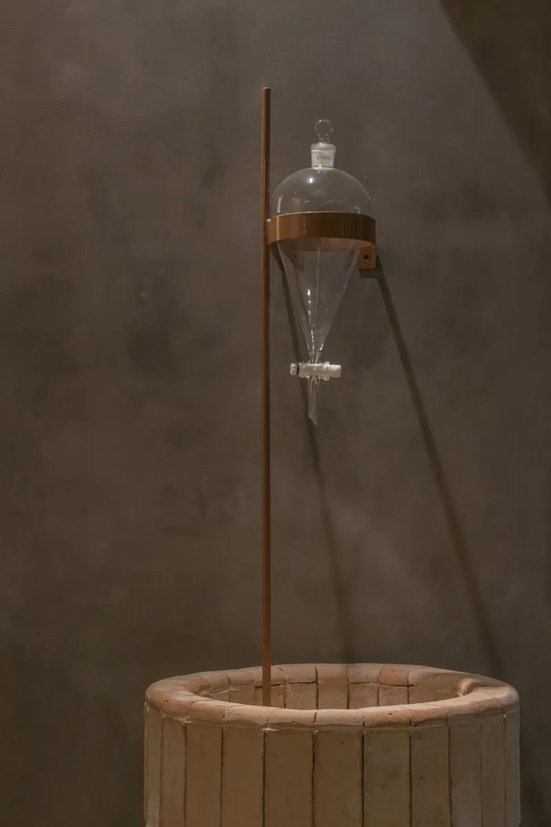 Glass separatory funnel suspended over a circular timber basin on a copper stand against plaster wall