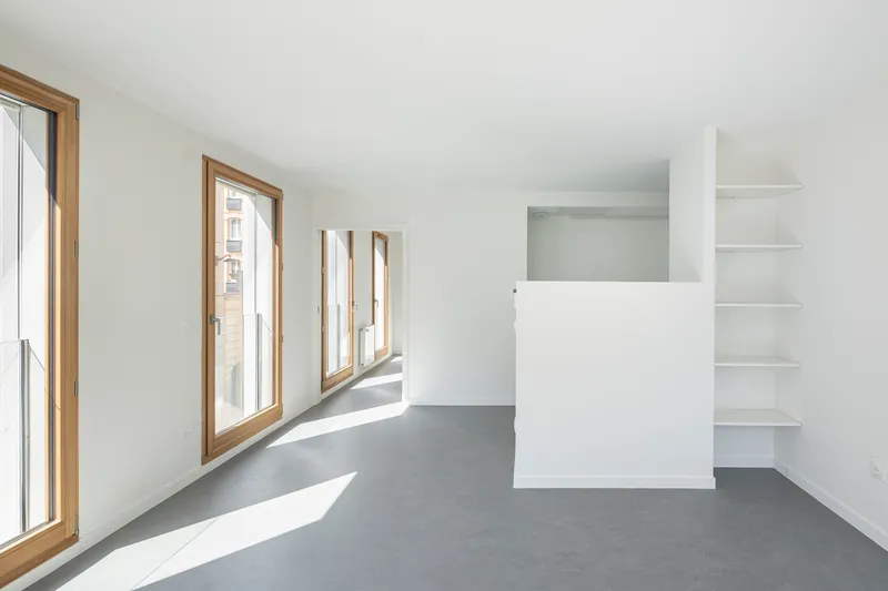 Open room with timber-framed windows casting shadows across grey flooring and white built-in shelving