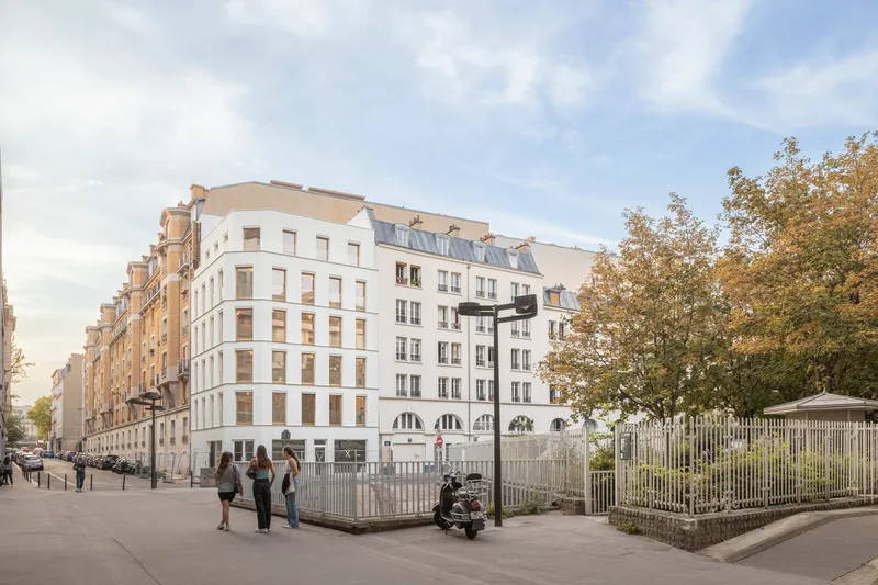 Street view of white residential block with pedestrians walking past white picket fence and mature trees in autumn
