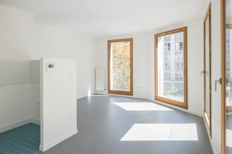 Open plan interior with grey floor, white walls, timber casement windows, and sunlight patches