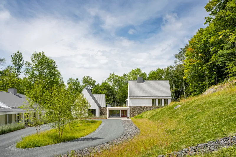 Cluster of white metal-clad gabled volumes along a curving gravel drive cutting through a grassy hillside