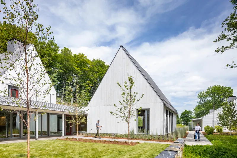 White vertical-board gable building with young trees in a planted lawn under partly cloudy skies