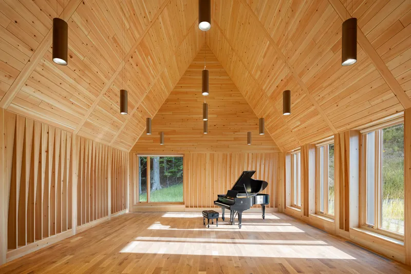 Interior of the timber-clad gable hall with pendant cylinders and a grand piano in sunlight