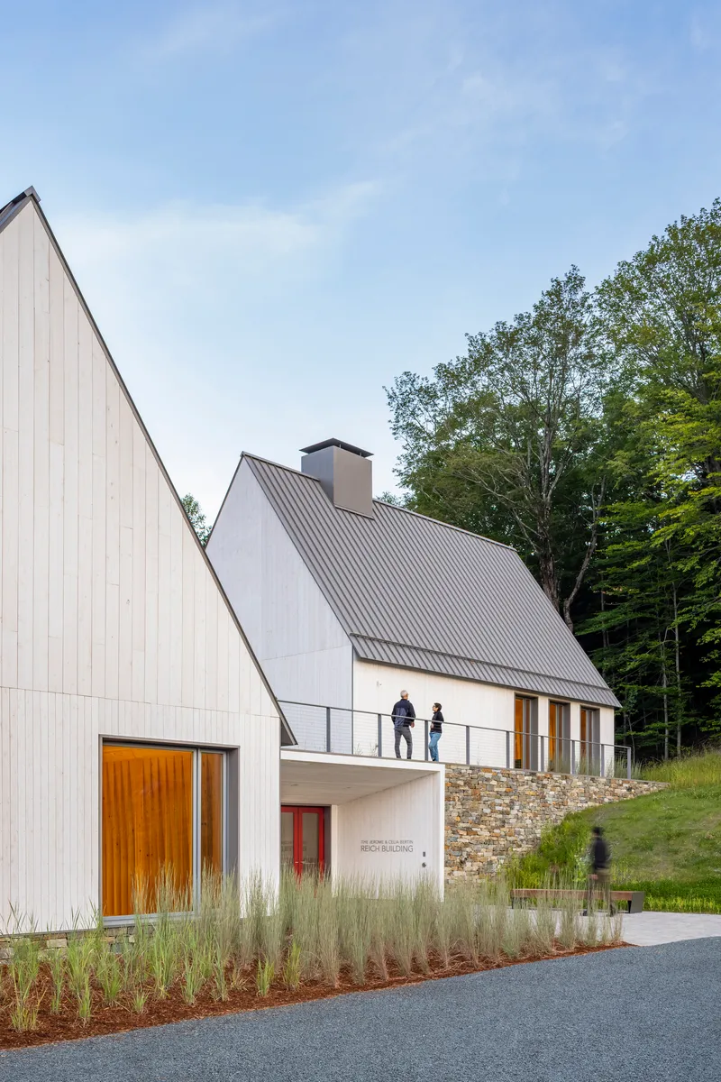 White vertical board facade with balcony and planted grasses along the gravel drive at dusk
