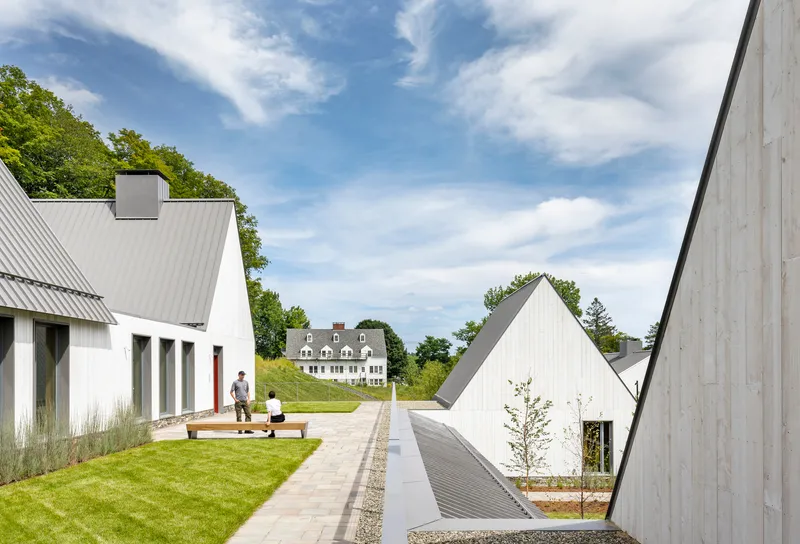 Central paved pathway between gabled volumes with two figures on a lawn under scattered clouds