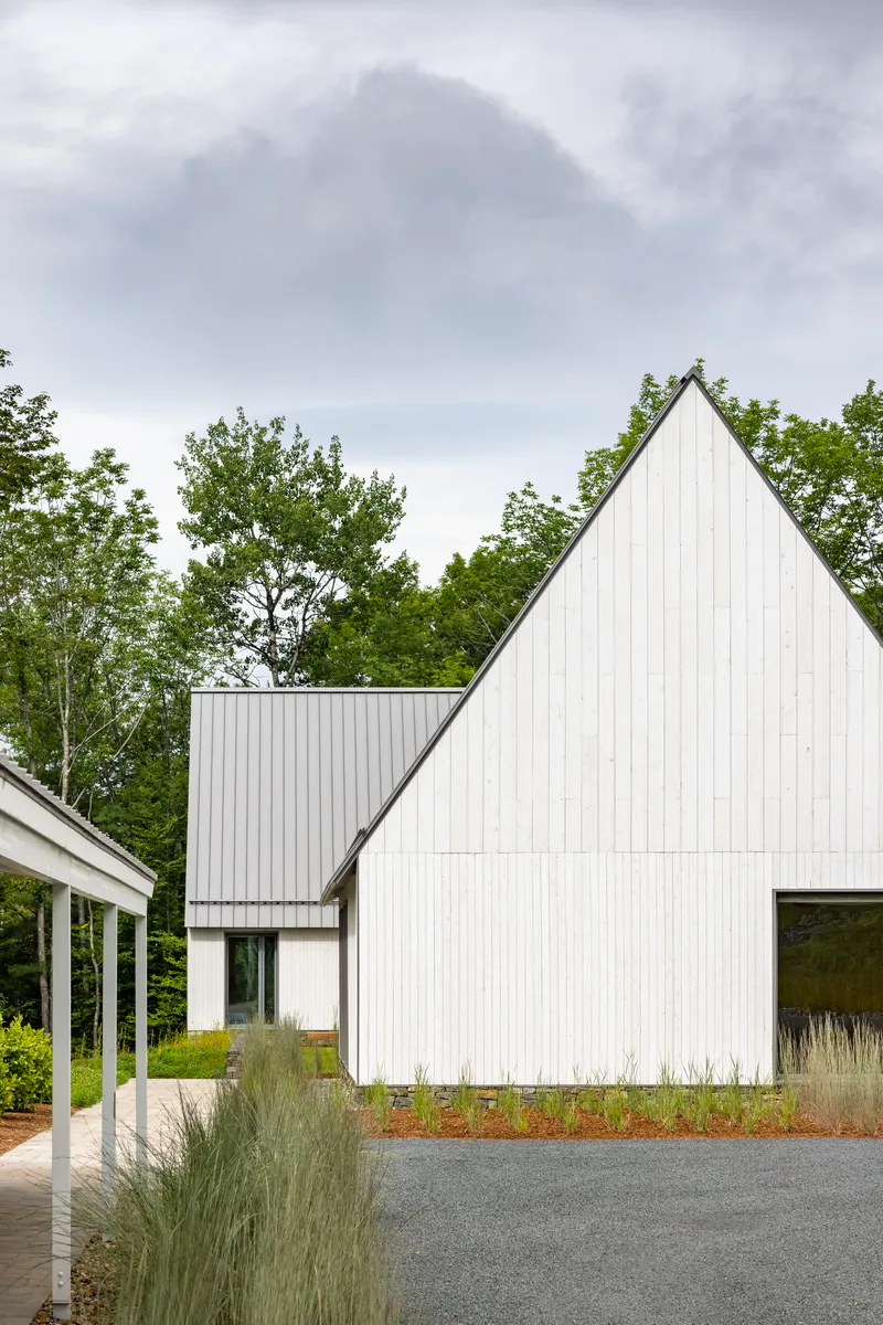 White cladded gable end with planted grasses beside a gravel drive under cloudy skies