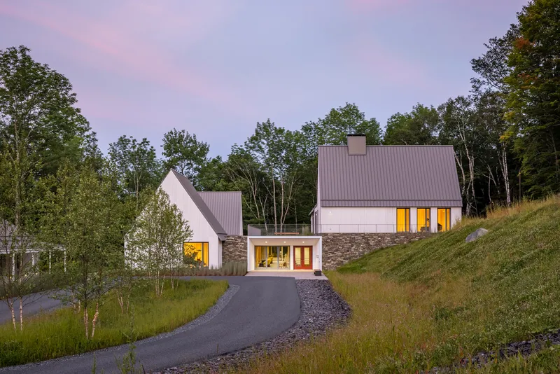 Cluster of gabled volumes with stone bases and glowing windows along a sloping drive at twilight