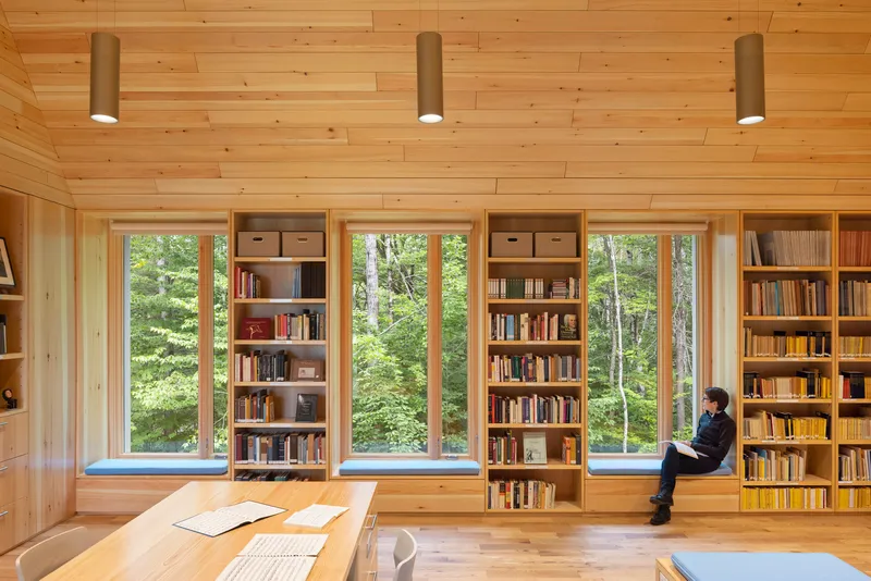 Light timber library with built-in bookshelves and tall windows overlooking summer foliage, person reading in windowsill