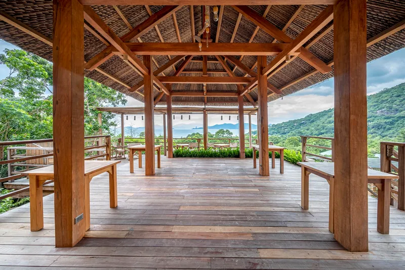 Open-air pavilion with exposed timber frame and thatched roof overlooking green valley