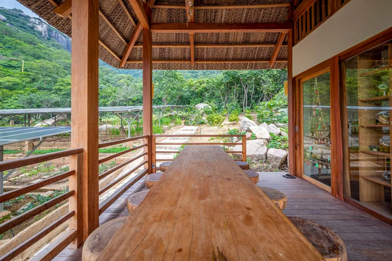 Covered terrace with woven bamboo ceiling and timber columns framing a live-edge table overlooking green hills