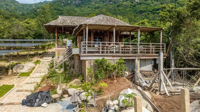 Elevated timber pavilion with thatched roofs and horizontal railings above a terraced garden in progress