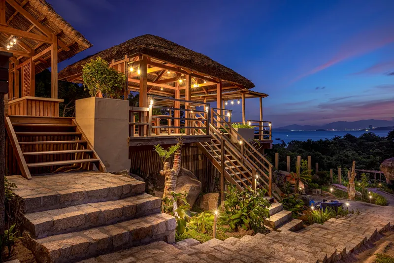 Stone staircase leading to the raised thatched pavilion illuminated at dusk with city lights beyond