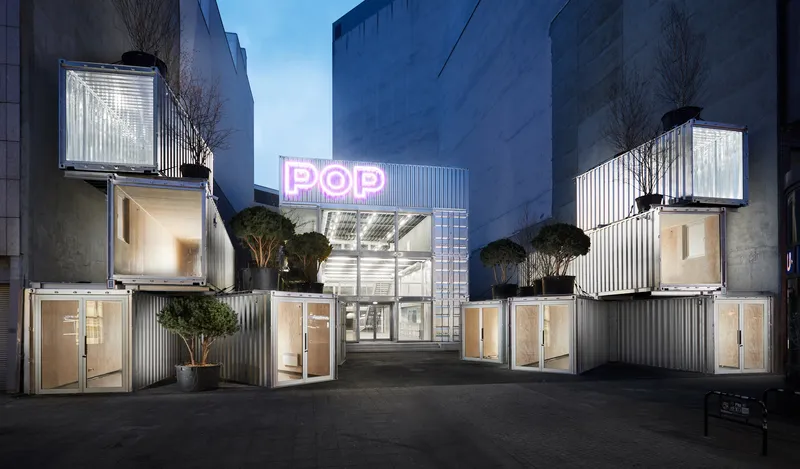 Courtyard view of stacked corrugated metal containers with planted trees and a neon sign at dusk