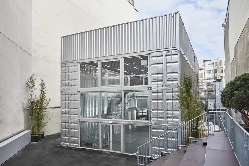Three-story corrugated metal structure with glass openings and an external metal staircase under daylight