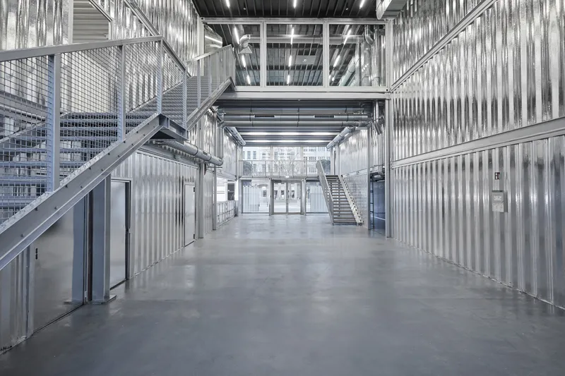 Interior hallway with corrugated metal walls, exposed ceiling ducts, and metal staircases flanking the passage