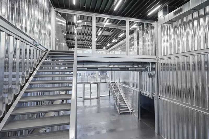 Steel staircase ascending between translucent corrugated walls beneath black ceiling with linear lighting strips